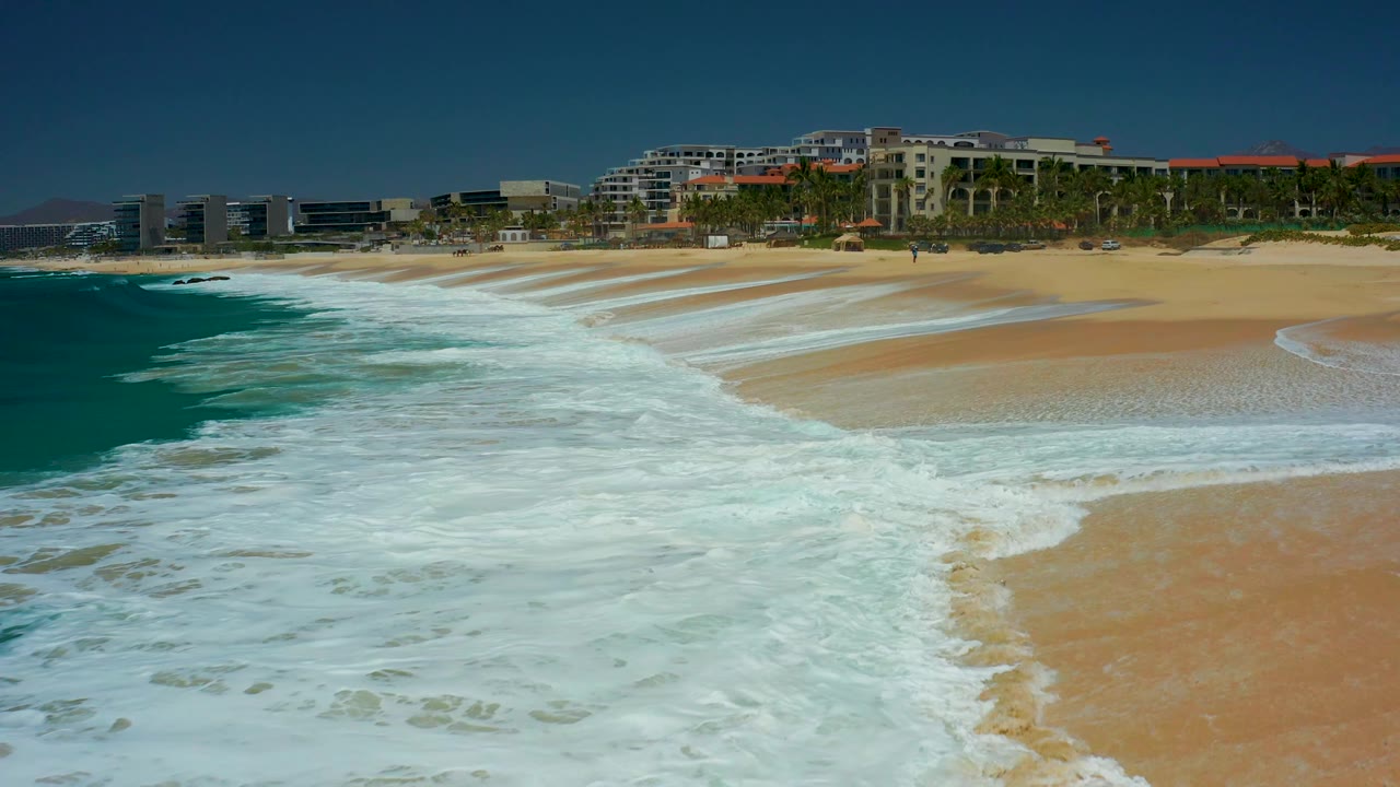 At 0:03, broad whitewater covers the lower beach while a darker turquoise wave edge approaches from the left, with Dreams Los Cabos buildings and palm trees across the background and no visible people.