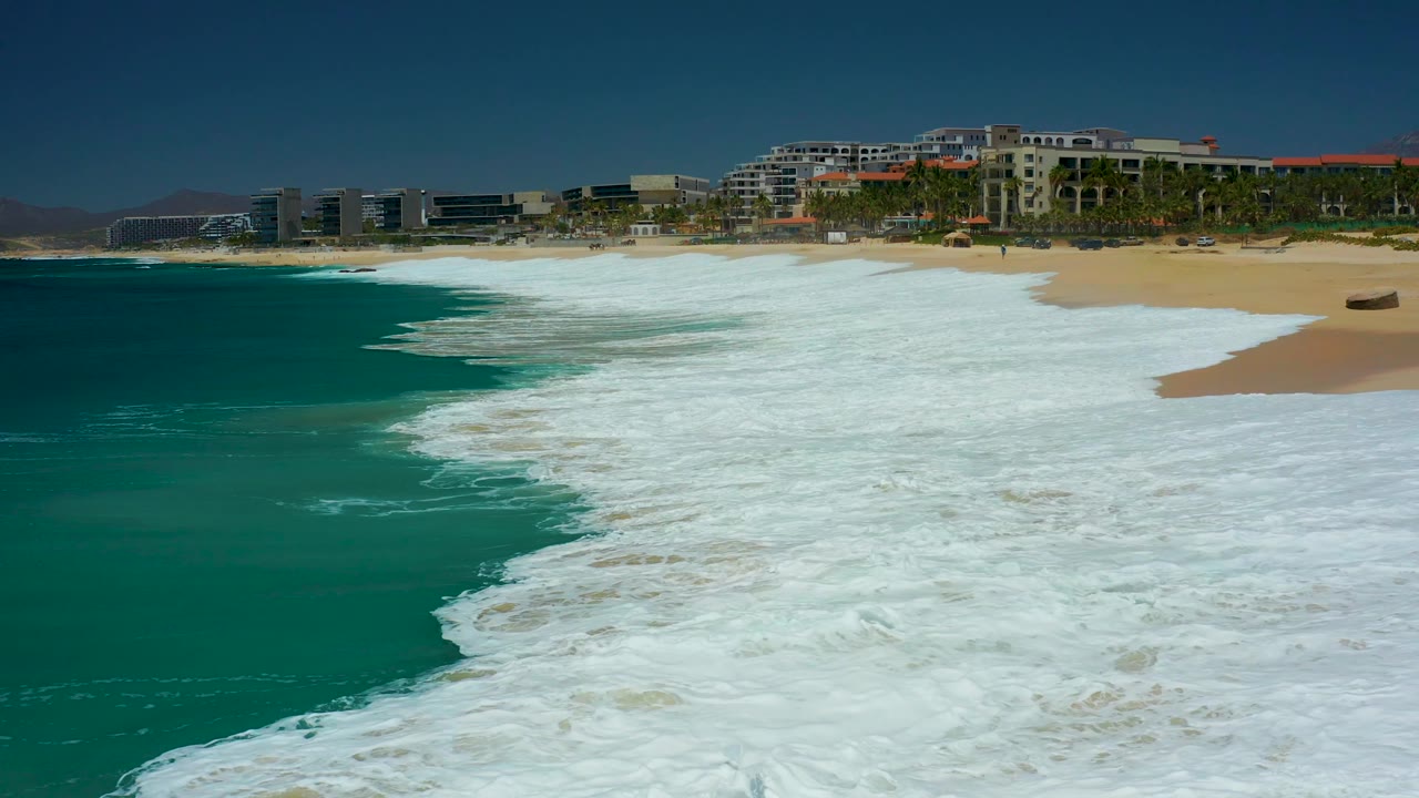 At Dreams Los Cabos, dense white surf covers much of the shoreline while a strip of tan beach and resort frontage remains visible along the top of the frame, with no visible people.