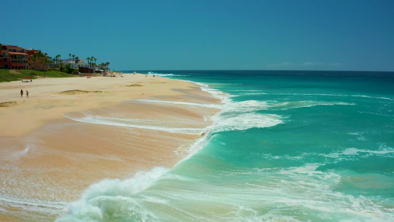 Aerial view at Dreams Los Cabos, Mexico, showing turquoise water curling onto pale sand, with small distant people on the left beach and resort buildings beyond.