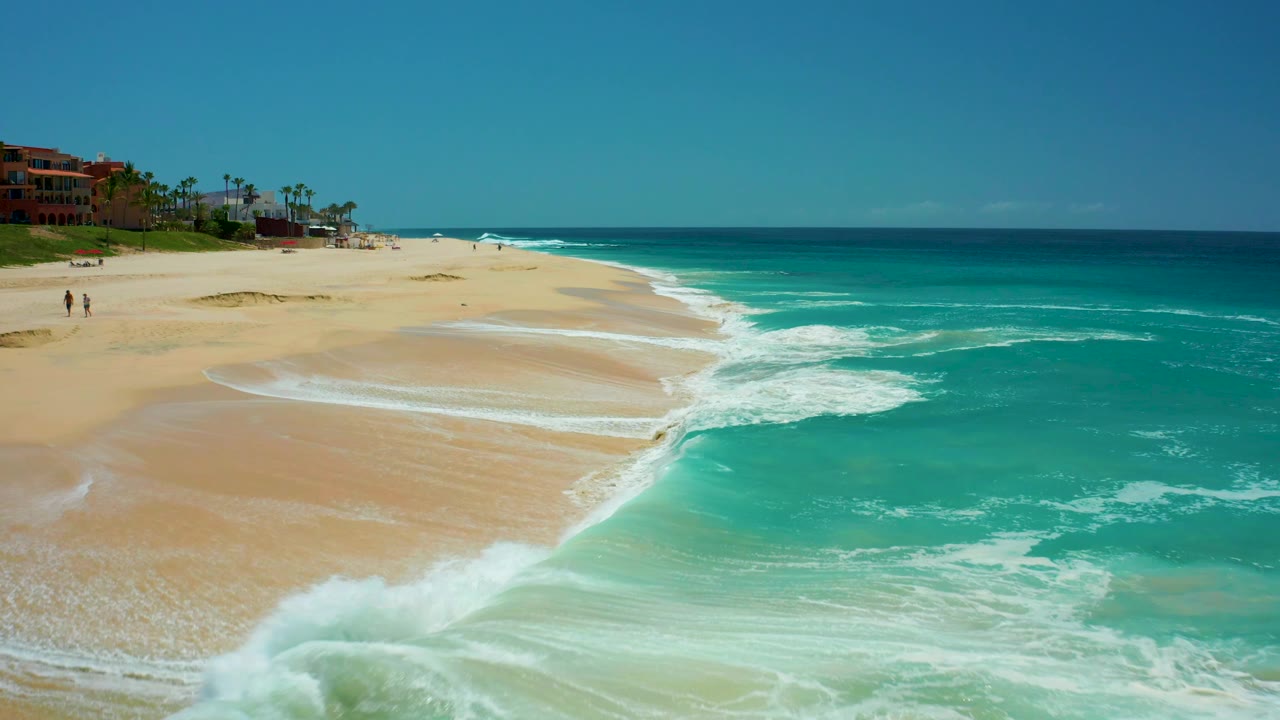 Turquoise surf spreads across a sandy shoreline below distant beach buildings.