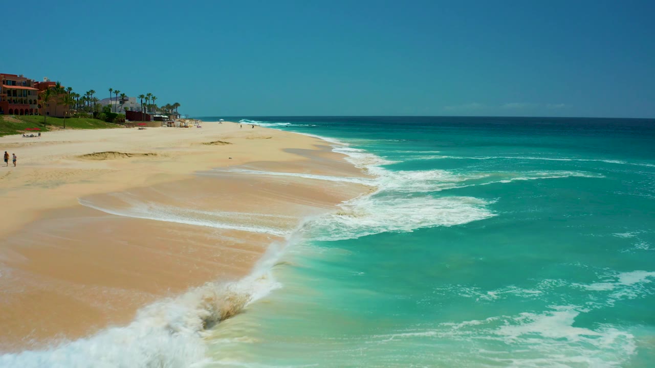 At Dreams Los Cabos, a larger breaking wave throws white foam across the lower shoreline while the turquoise water fills the right side and distant beachgoers remain small at left.