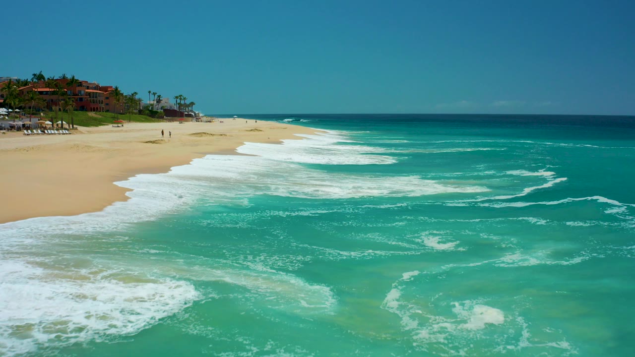 At Dreams Los Cabos in Baja California Sur, turquoise surf spreads white foam over light sand with resort buildings and palm trees in the upper left and tiny distant beachgoers on the beach.