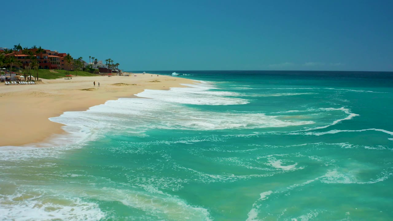 The shoreline at Dreams Los Cabos is seen from above with more surf filling the right side, pale sand on the left, tiny beachgoers near the upper beach, and a clear ocean horizon.