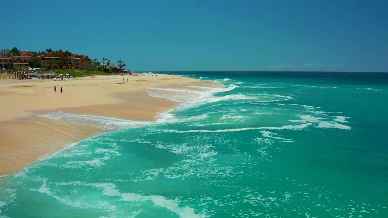 Opening frame of turquoise surf rolling onto the sandy beach at Dreams Los Cabos, with distant resort buildings, palms, blue sky, and small people on the sand.