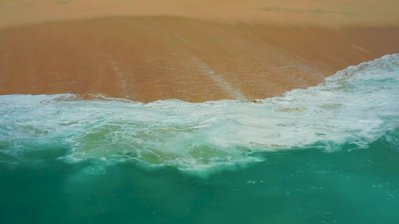 Aerial shoreline view at Dreams Los Cabos with a broad white foam line spreading over the tan beach edge, no visible people.