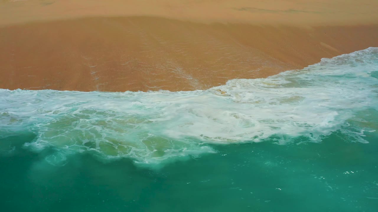 Top-down frame at Dreams Los Cabos showing turquoise-green water below a foamy shore break and smooth tan sand above, no visible people.