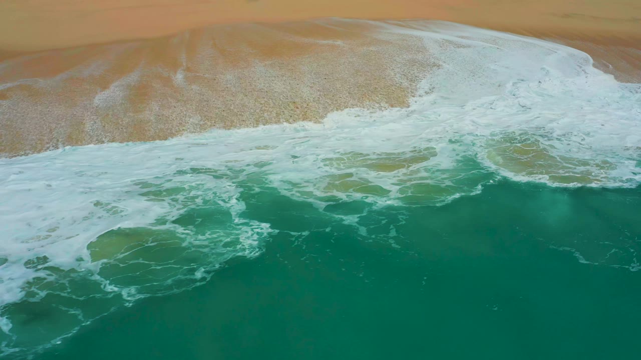 Aerial shoreline view at Dreams Los Cabos with the foamy wave edge thinning across wet sand and green water filling the lower frame, no visible people.