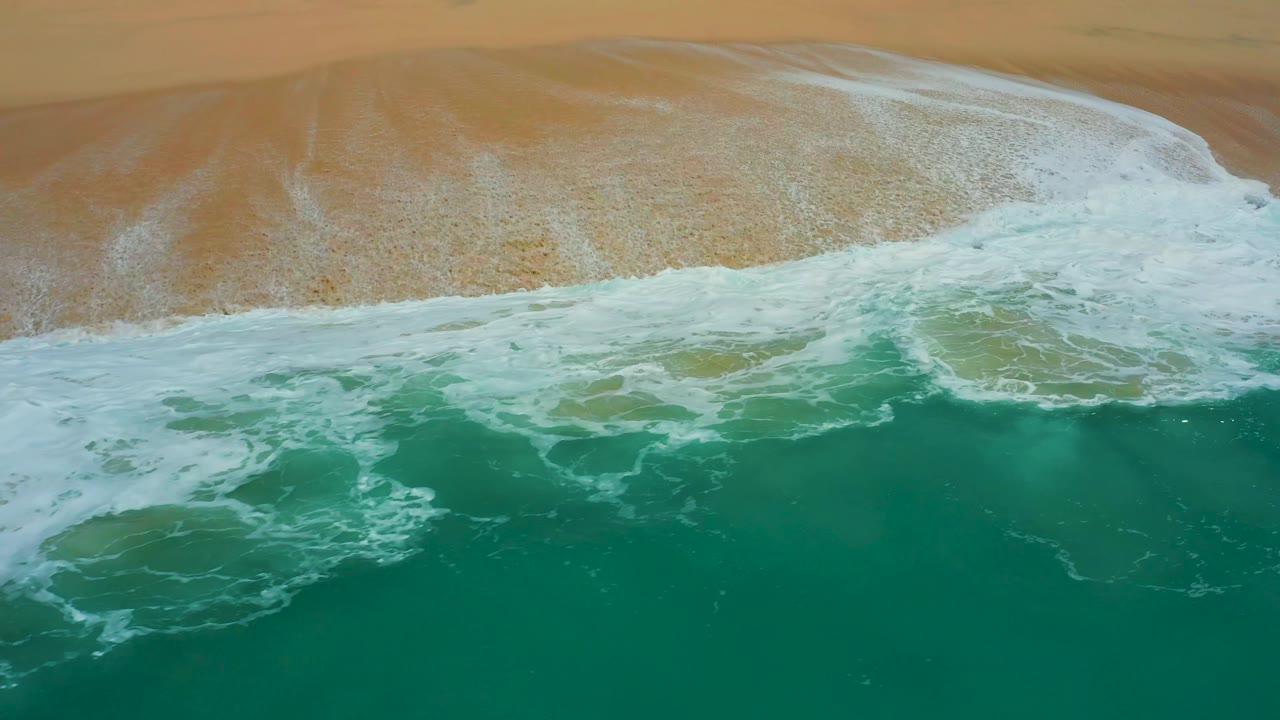 Top-down beach frame from Dreams Los Cabos showing a curved white surf line between broad tan sand and darker green ocean, no visible people.