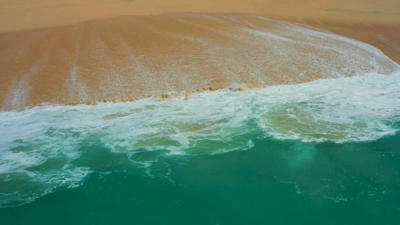 Overhead Dreams Los Cabos beach view with more exposed tan sand and trailing white foam patterns near the lower edge, no visible people.