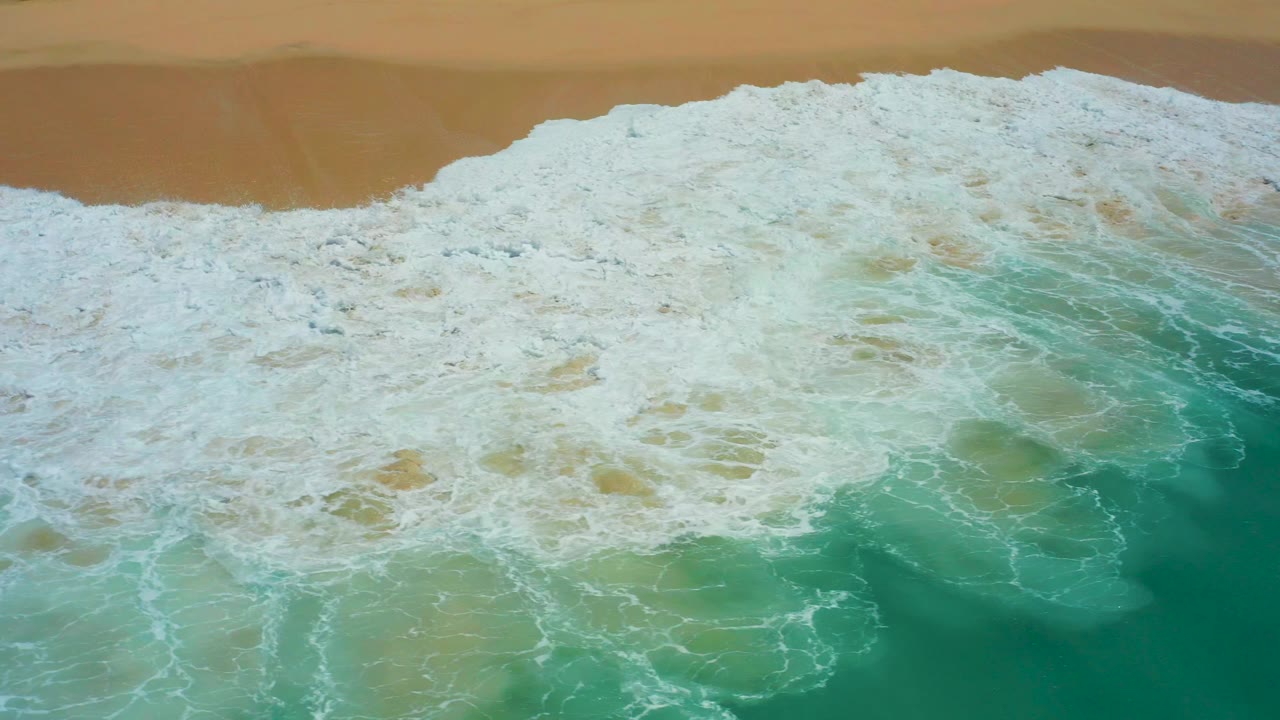 At Dreams Los Cabos in Baja California Sur, white foam spreads across turquoise water toward a tan sandy beach, with no visible people.