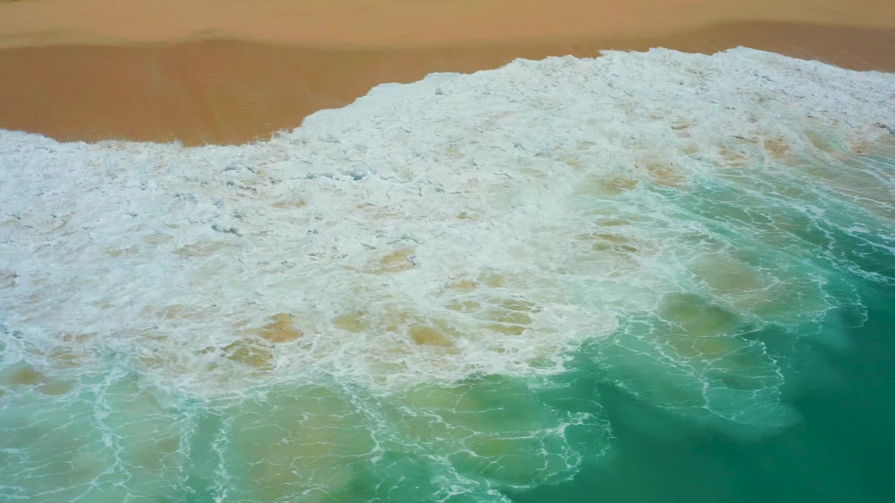 A high aerial view shows a broad foamy wave band moving across green-blue water toward the sand at Dreams Los Cabos, with no visible people.