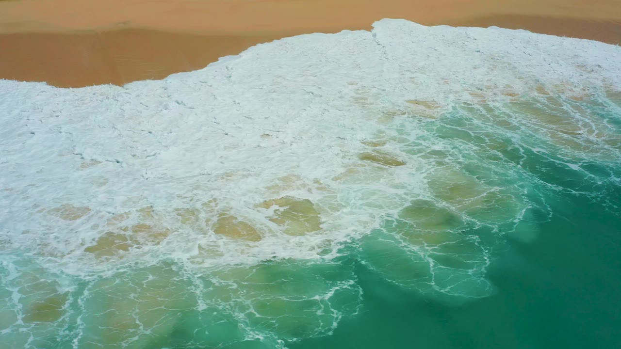 Whitewater fills much of the frame beside a narrow strip of tan beach at Dreams Los Cabos, while deeper turquoise water remains visible at the lower right, with no visible people.