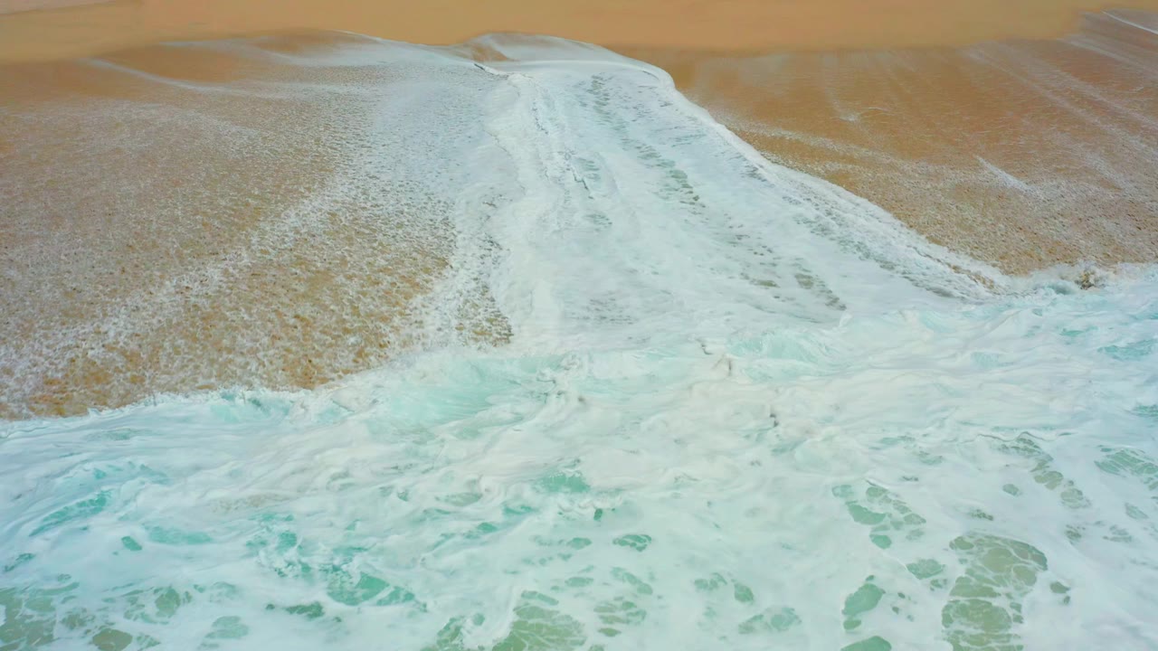 Aerial view at Dreams Los Cabos, Mexico, showing white surf fanning over tan beach sand with turquoise water at the lower edge and no visible people.