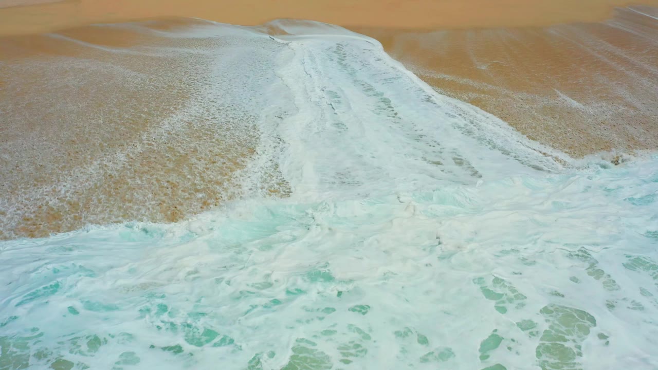 A top-down shoreline frame at Dreams Los Cabos, Mexico, with pale foam covering the center of the beach and a strip of dry tan sand above, with no visible people.