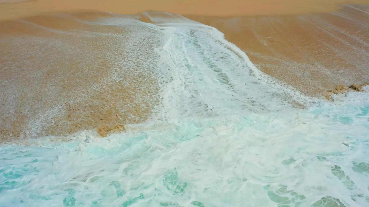 Aerial beach view at Dreams Los Cabos, Mexico, where white surf cuts diagonally across tan sand and turquoise water gathers along the lower frame, with no visible people.