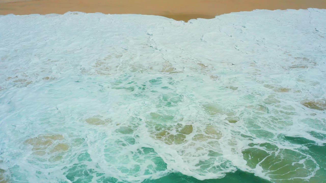 Overhead surf foam and turquoise water meeting tan sand.