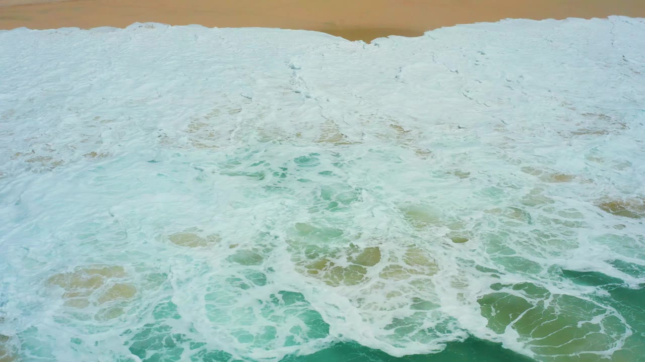 At 0:01, the foamy wave field thickens across the center of the Dreams Los Cabos shoreline frame, with green water visible through broken foam and no visible people.