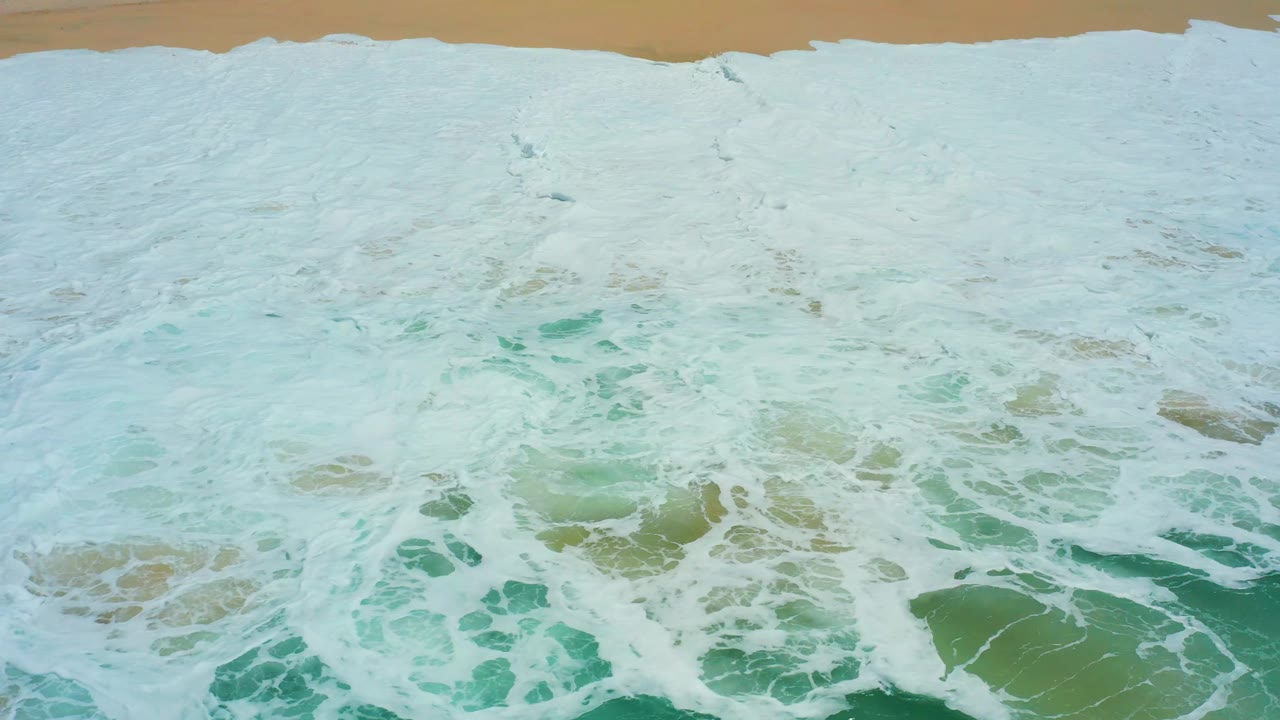 At 0:02, a broad white breaker pushes toward the top sand line at Dreams Los Cabos while darker turquoise water pools near the lower right and no visible people appear.