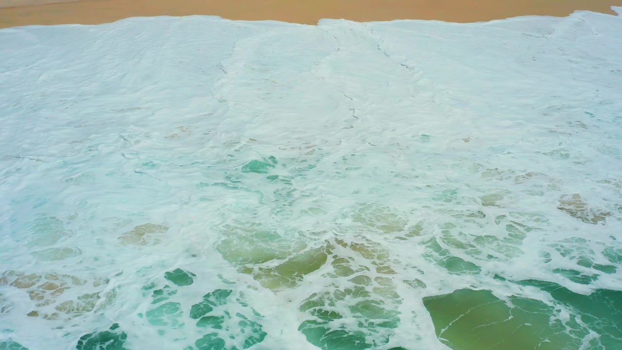 At 0:03, pale foam nearly blankets the overhead ocean view at Dreams Los Cabos, leaving a narrow strip of tan sand along the top and no visible people.