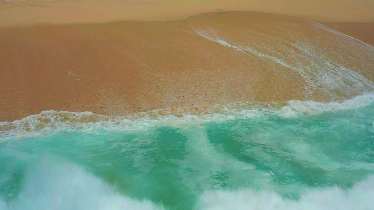 Aerial frame from Dreams Los Cabos with turquoise water pressing closer to the sand as a wider white foam line forms along the shore, with no visible people.