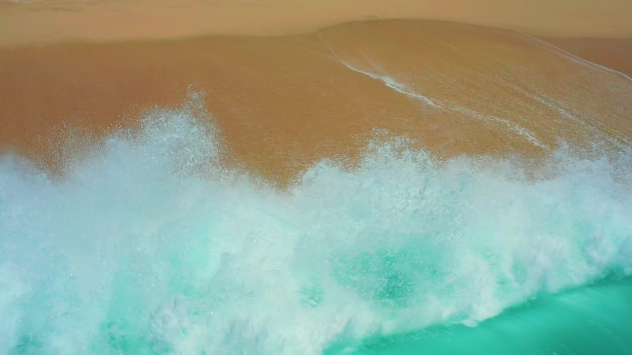 Top-down Dreams Los Cabos surf view where a large white breaking wave pushes into the turquoise water below the tan beach, with no visible people.