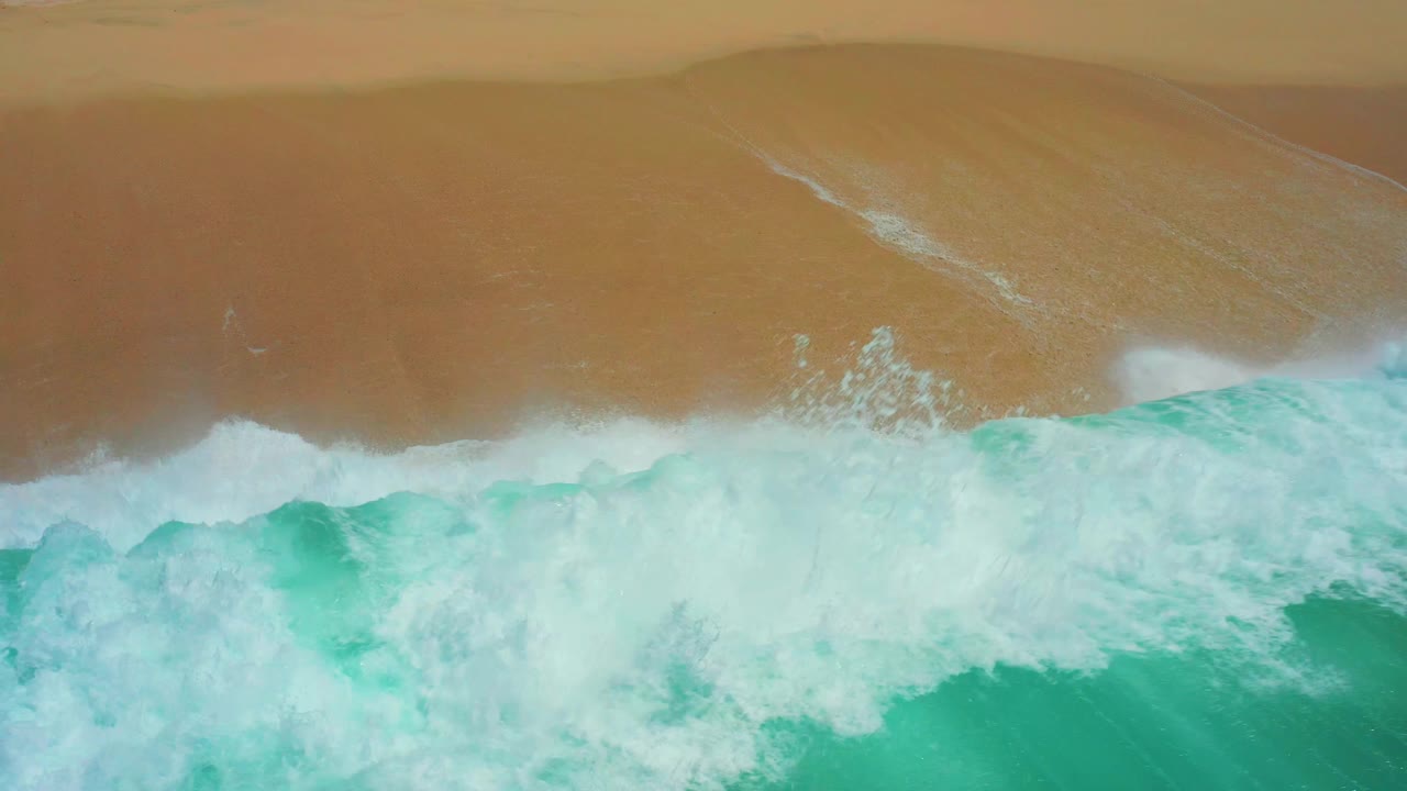 Overhead beach frame at Dreams Los Cabos showing white surf spreading broadly across the shoreline and covering much of the turquoise water, with no visible people.