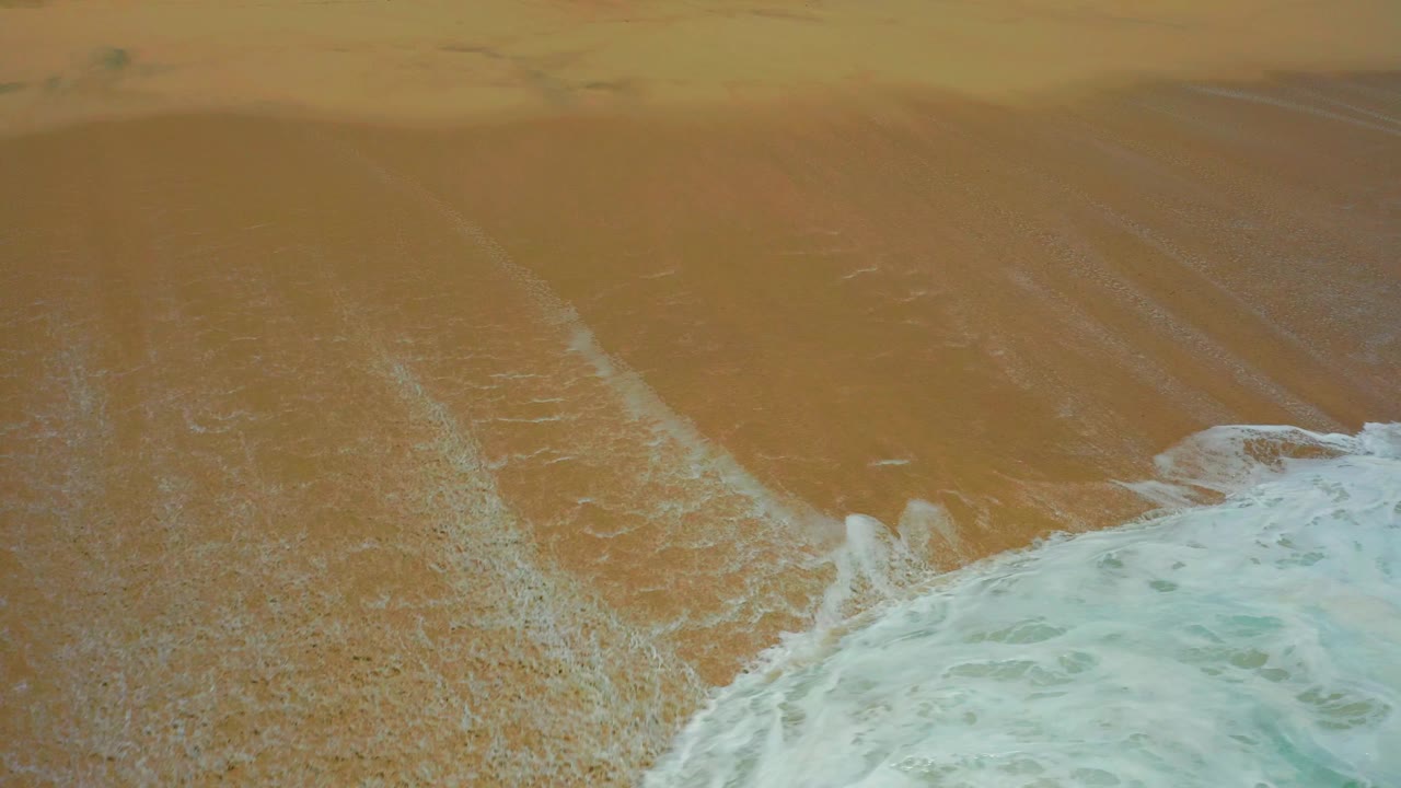 Foamy ocean water spreads farther along the lower right of the Dreams Los Cabos shoreline, leaving thin pale trails on the wet sand and no visible people.