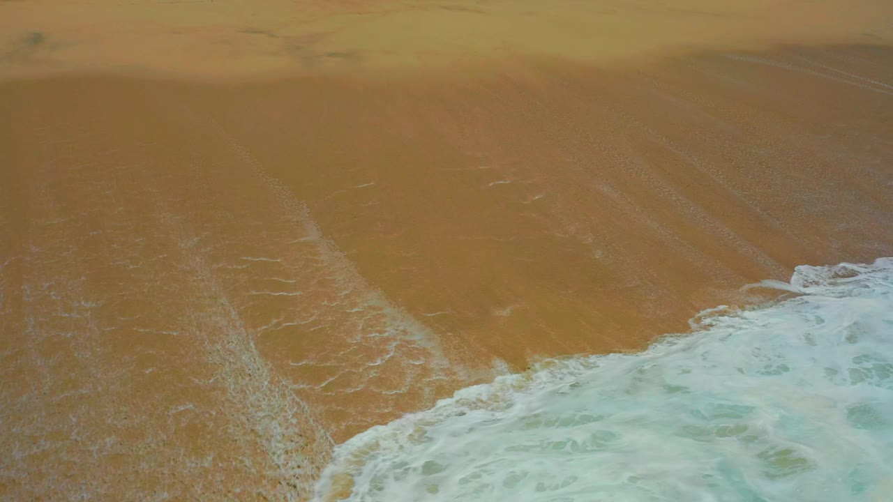 A turquoise wave edge crosses the bottom right of the frame while smooth golden sand fills the upper area at Dreams Los Cabos, with no visible people.