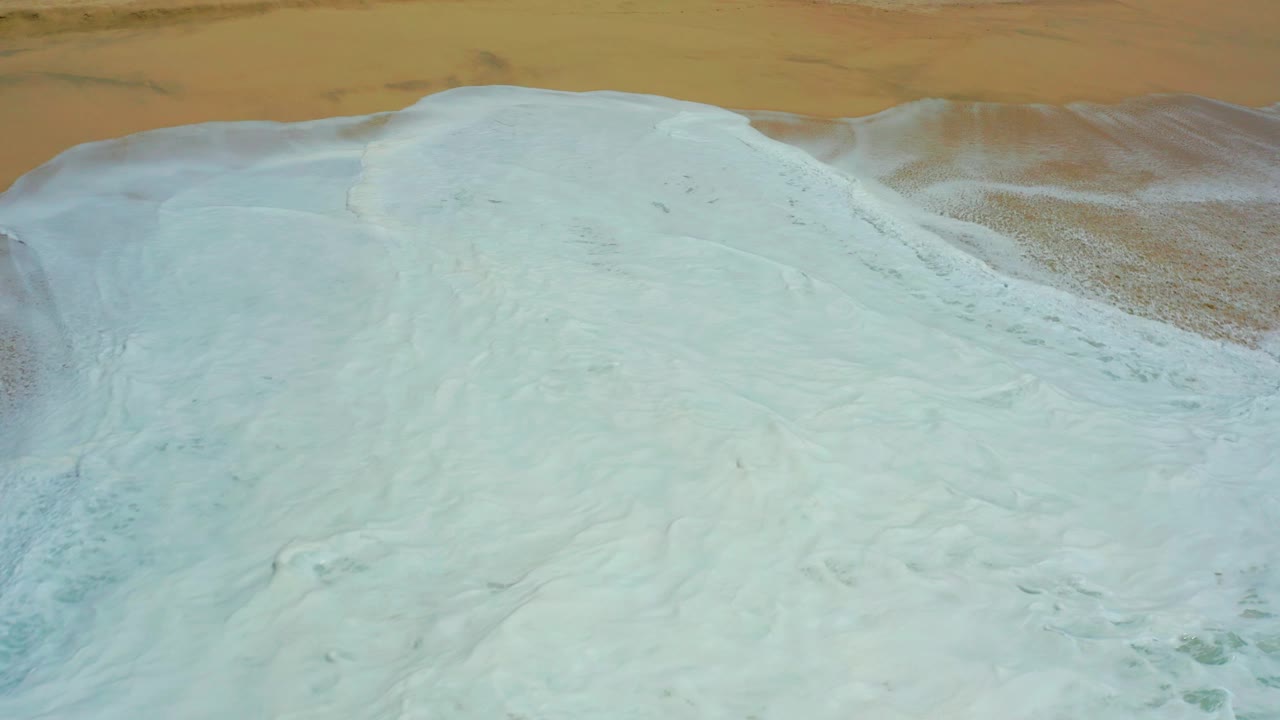 Aerial view at Dreams Los Cabos showing white ocean foam covering most of the tan sandy shore, with no visible people.