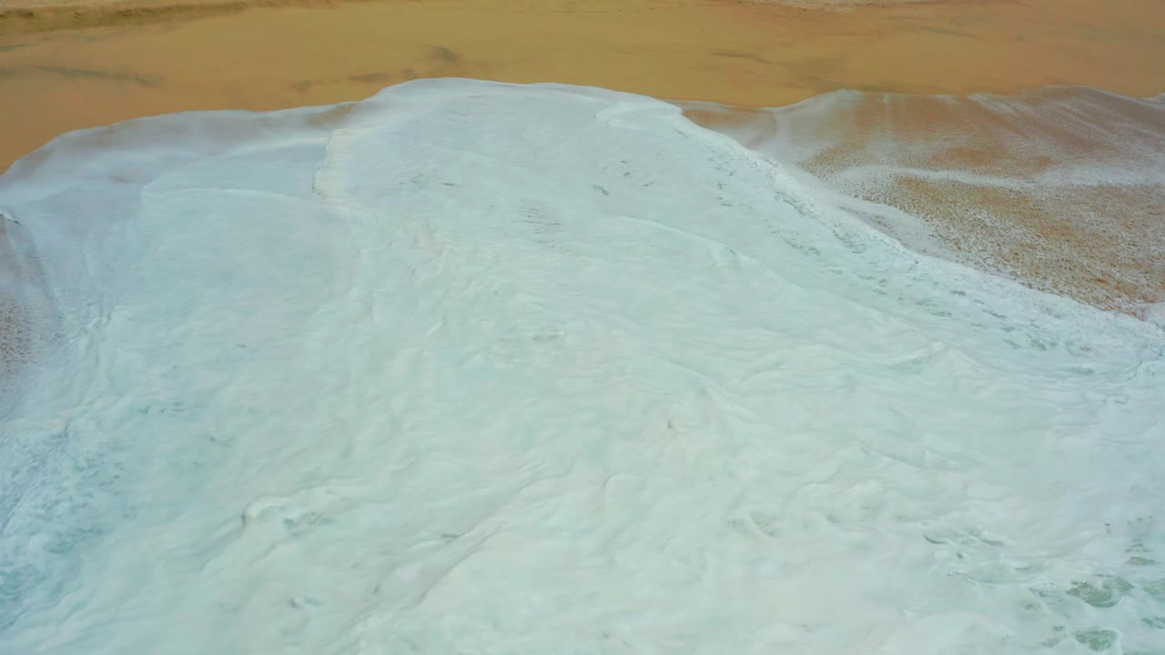 Aerial frame from Dreams Los Cabos with pale foam still blanketing the shoreline and thin tan sand visible along the top edge, with no visible people.
