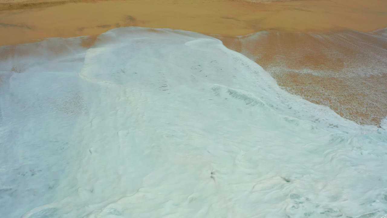 Overhead view at Dreams Los Cabos where the wave foam forms a rounded pale mass against tan beach sand, with no visible people.