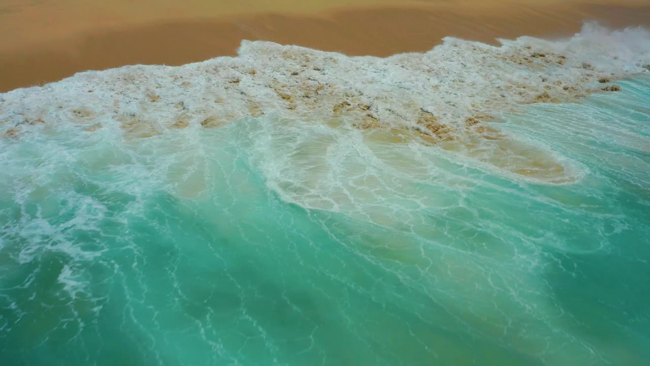 Aerial frame from Dreams Los Cabos where the foamy wave front spreads farther across the turquoise water and reaches the sand, with no visible people.