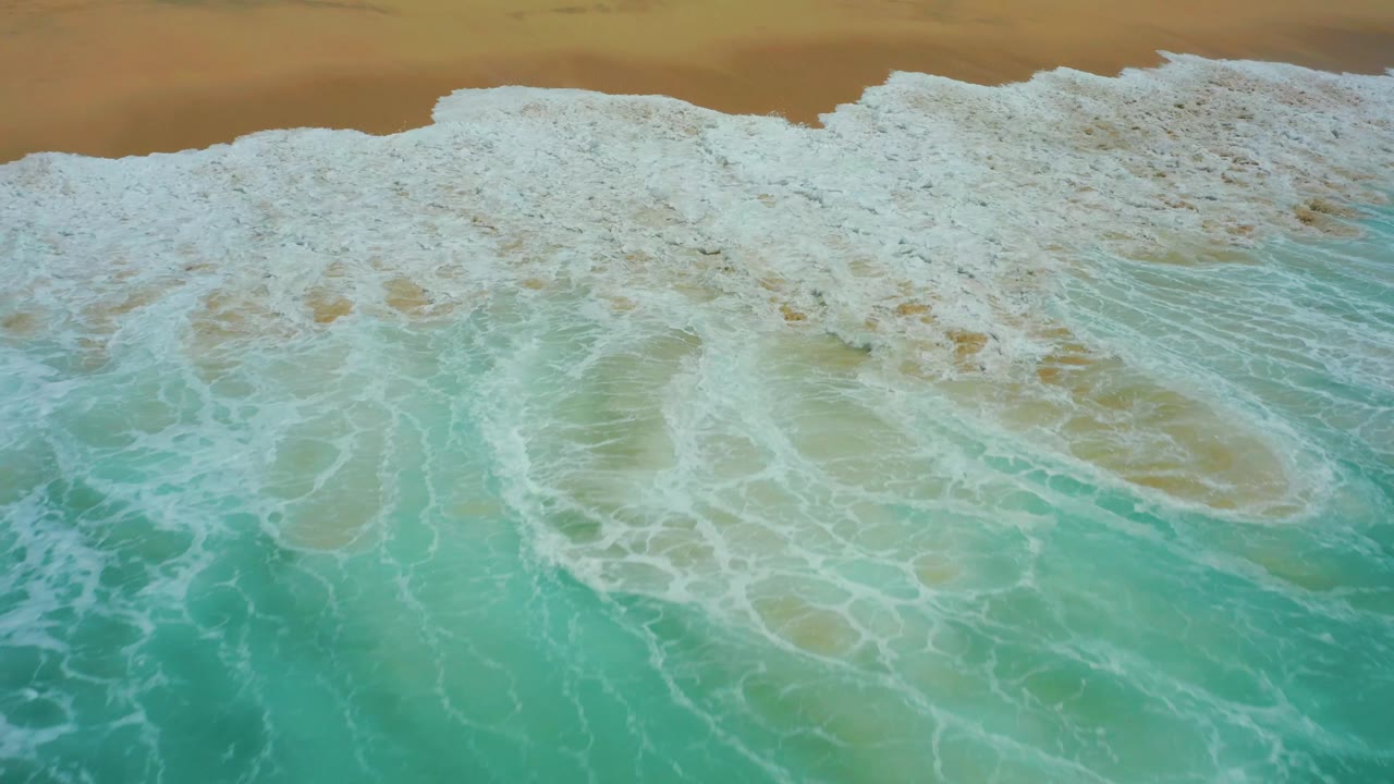Top-down Dreams Los Cabos shoreline frame with a larger sheet of white surf rolling over turquoise water below a narrow strip of tan beach, with no visible people.