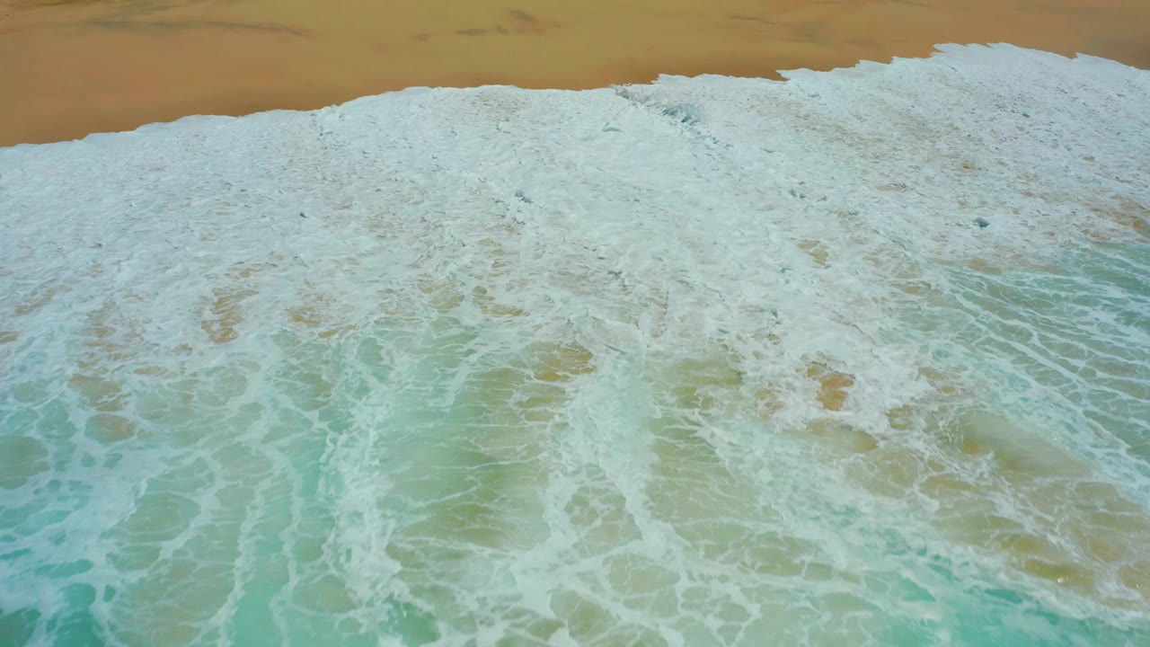 Aerial view at Dreams Los Cabos with white foam blanketing shallow turquoise water and a thin sandy beach band along the top, with no visible people.