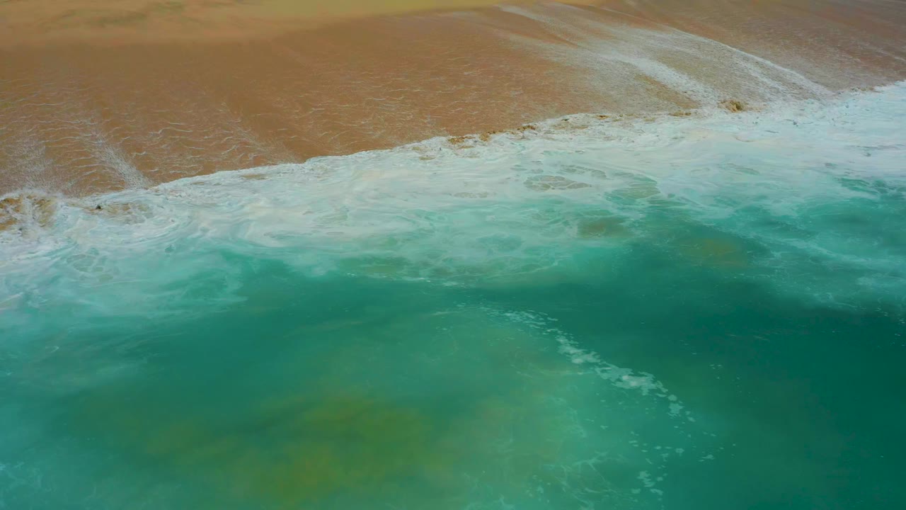 Overhead view at Dreams Los Cabos of turquoise water approaching a foamy line on tan sand, with no visible people.