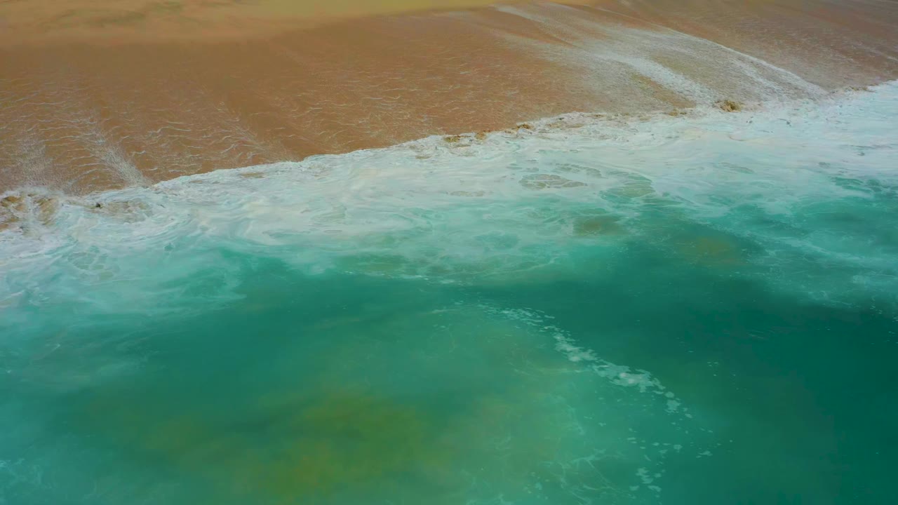Aerial frame of green-blue water and white foam spreading along the sandy beach at Dreams Los Cabos, with no visible people.