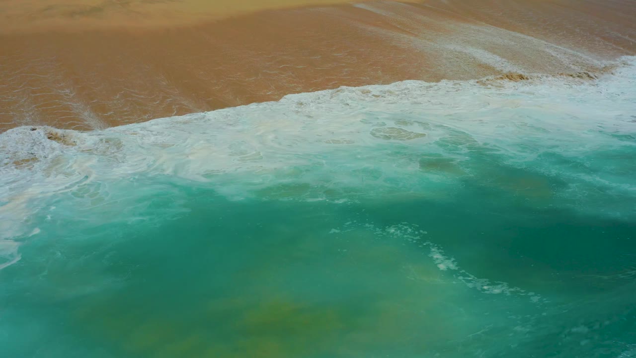 The wave edge turns white as it rolls diagonally onto the tan shore at Dreams Los Cabos, with no visible people.