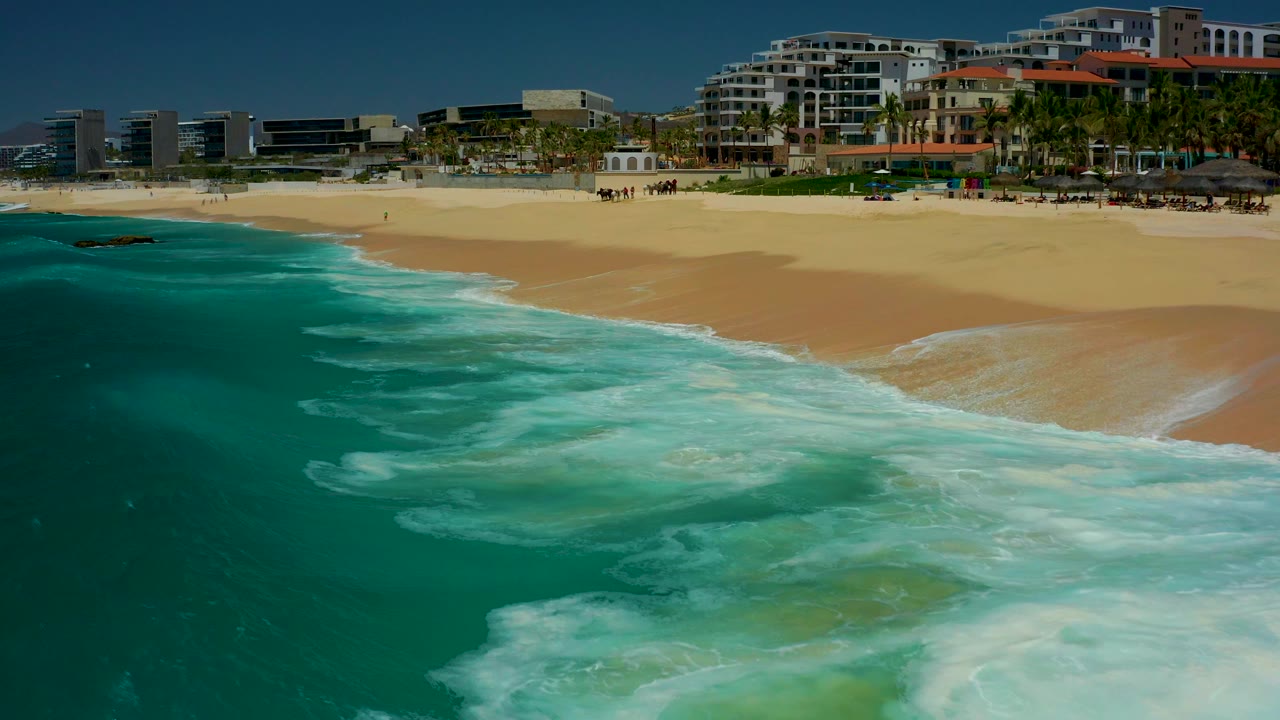 At Dreams Los Cabos in Baja California Sur, turquoise water and white foam approach a sandy beach while resort buildings and small distant people line the background.