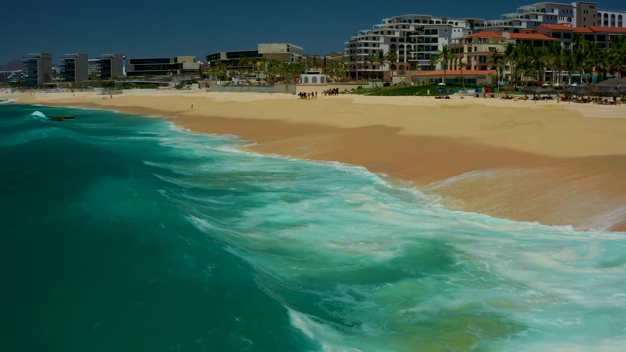 At Dreams Los Cabos in Baja California Sur, the wave breaks diagonally along the beach, leaving bright white foam between the turquoise water and the resort-lined shore.