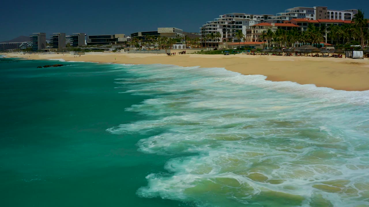 At Dreams Los Cabos in Baja California Sur, turquoise water and white foam approach the sandy beach while resort buildings and palms line the background, with no visible people.