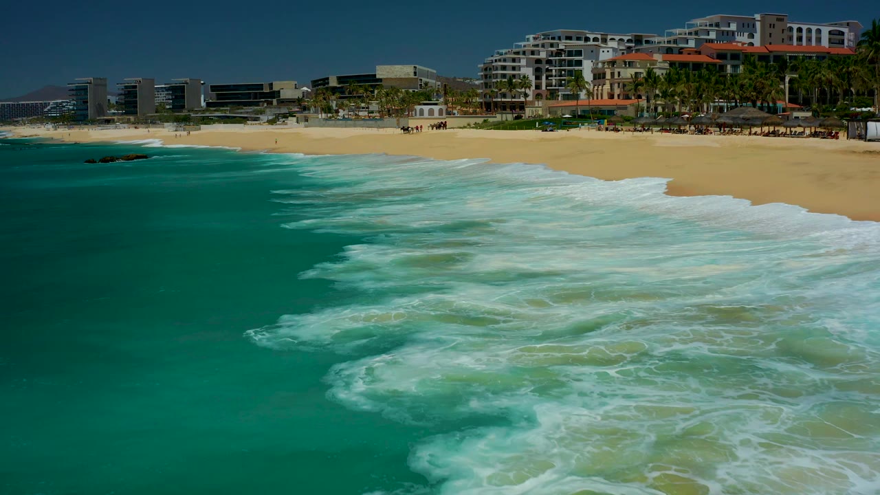 The wave line pushes closer to the Dreams Los Cabos beach, leaving white foam over shallow turquoise water with resort buildings and palms across the upper frame, with no visible people.
