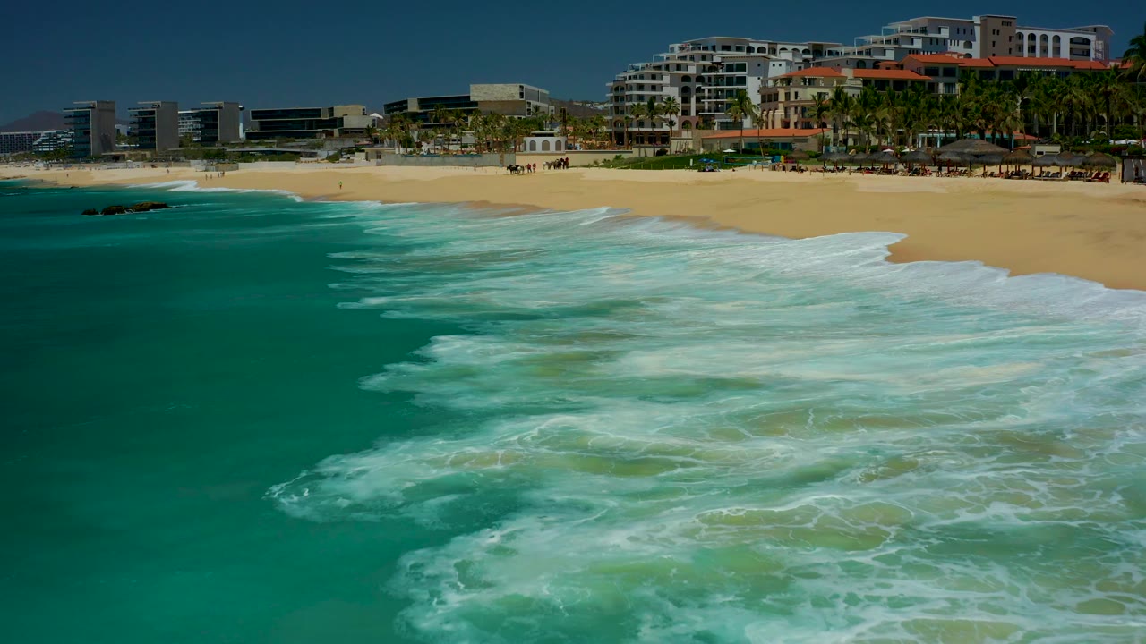 A broad sheet of white surf crosses turquoise water near a sandy resort beach.