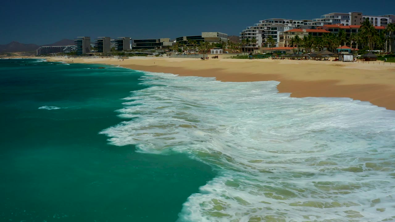 At 0:01, the foamy surf broadens across the lower frame while the sandy Dreams Los Cabos shoreline and beachfront buildings stay visible along the top, with no visible people.