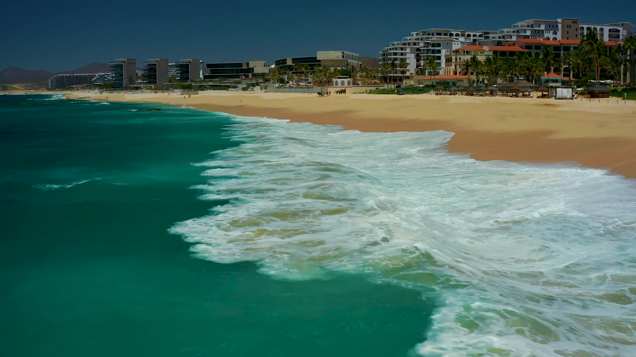 At 0:02, a larger band of white water curls diagonally across the turquoise ocean near the Dreams Los Cabos sand, backed by coastal buildings and no visible people.