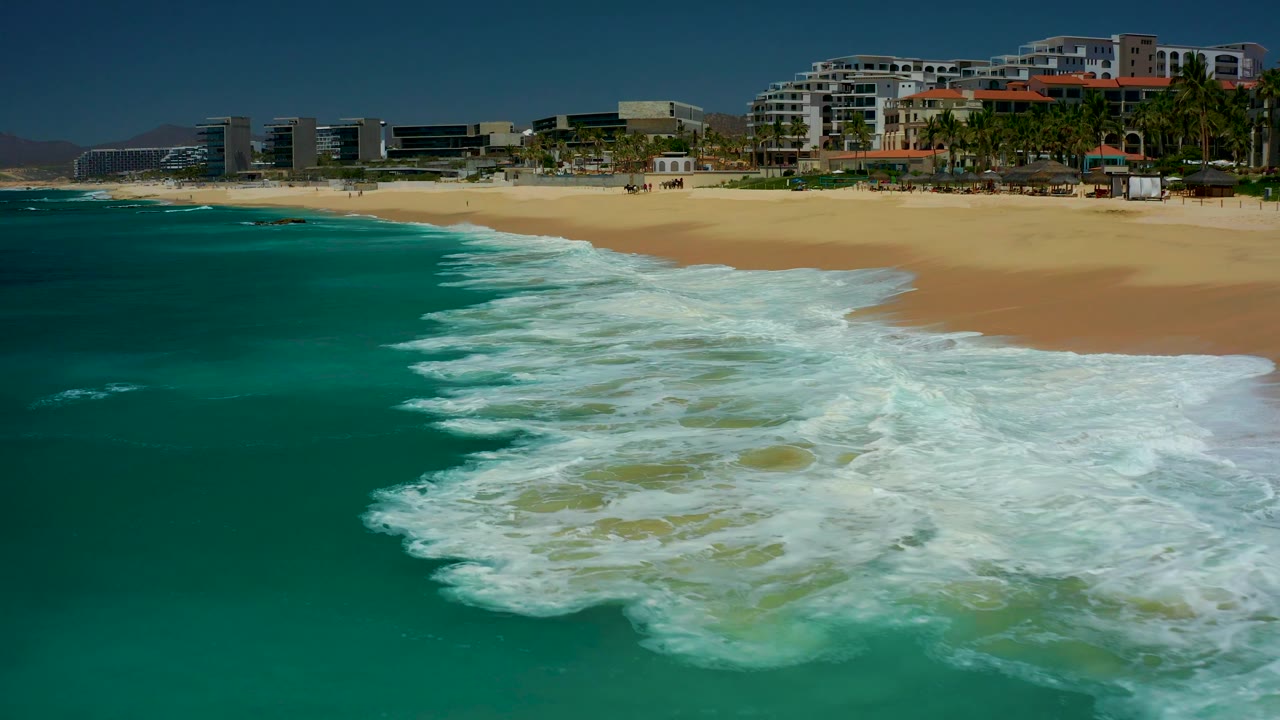 At 0:03, white foam covers more of the nearshore water while the pale sand and Dreams Los Cabos coastal buildings remain across the upper frame, with no visible people.