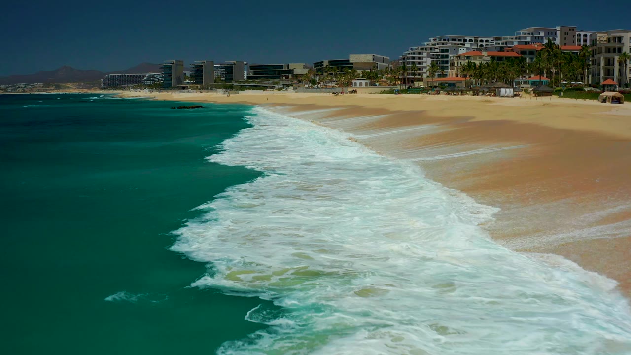 At 0:01, the foamy wave sheet advances along the diagonal shoreline at Dreams Los Cabos, with teal water on the left, dry sand on the right, and no visible people.