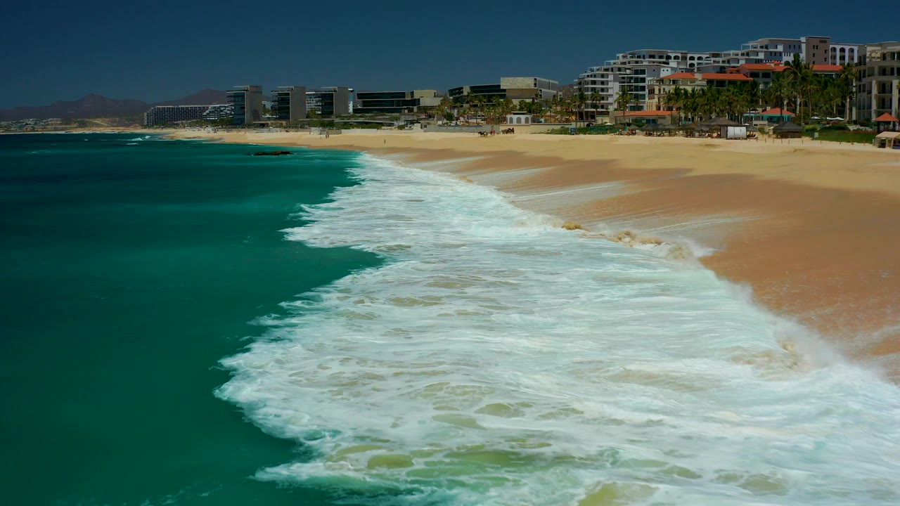 At Dreams Los Cabos, a larger band of white foam covers the lower shoreline while turquoise water remains open on the left and beachfront buildings line the upper right, with no visible people.