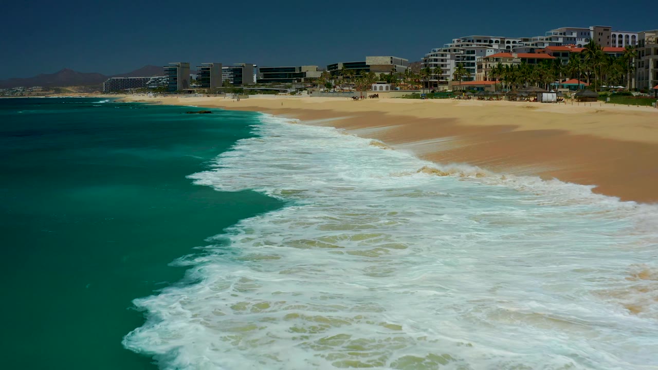 The 0:03 frame shows the whitewash stretched farther down the Dreams Los Cabos beach, with a clear split between deep teal water, pale sand, and no visible people.