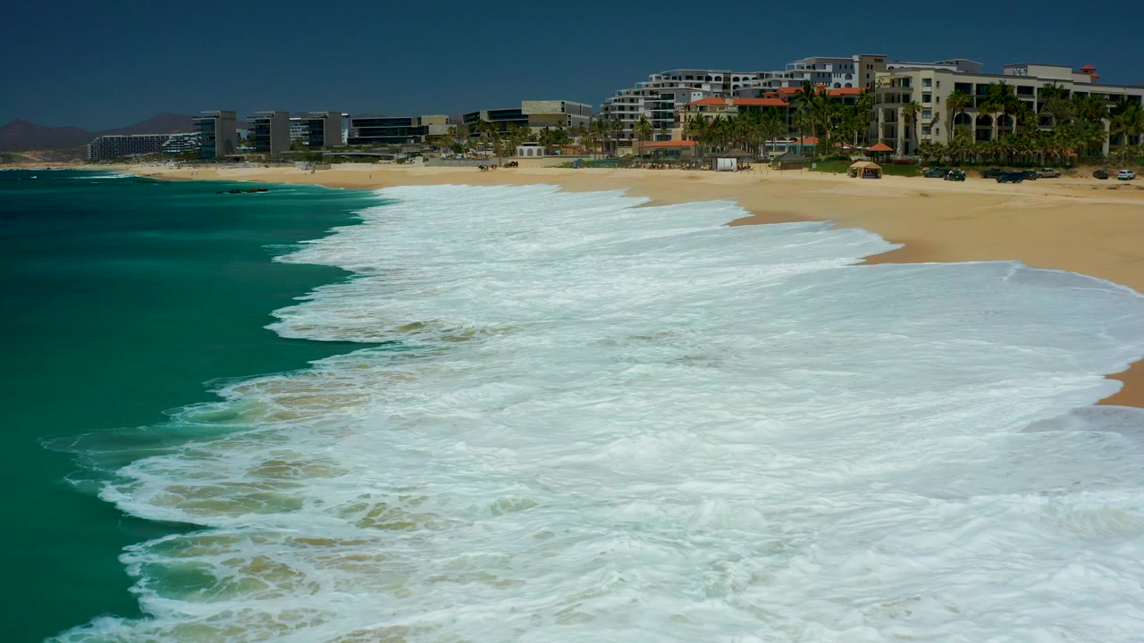 White foam pushes farther across the sandy shore at Dreams Los Cabos, with deep turquoise water on the left and a row of beachfront buildings and palms beyond the beach, with no visible people.