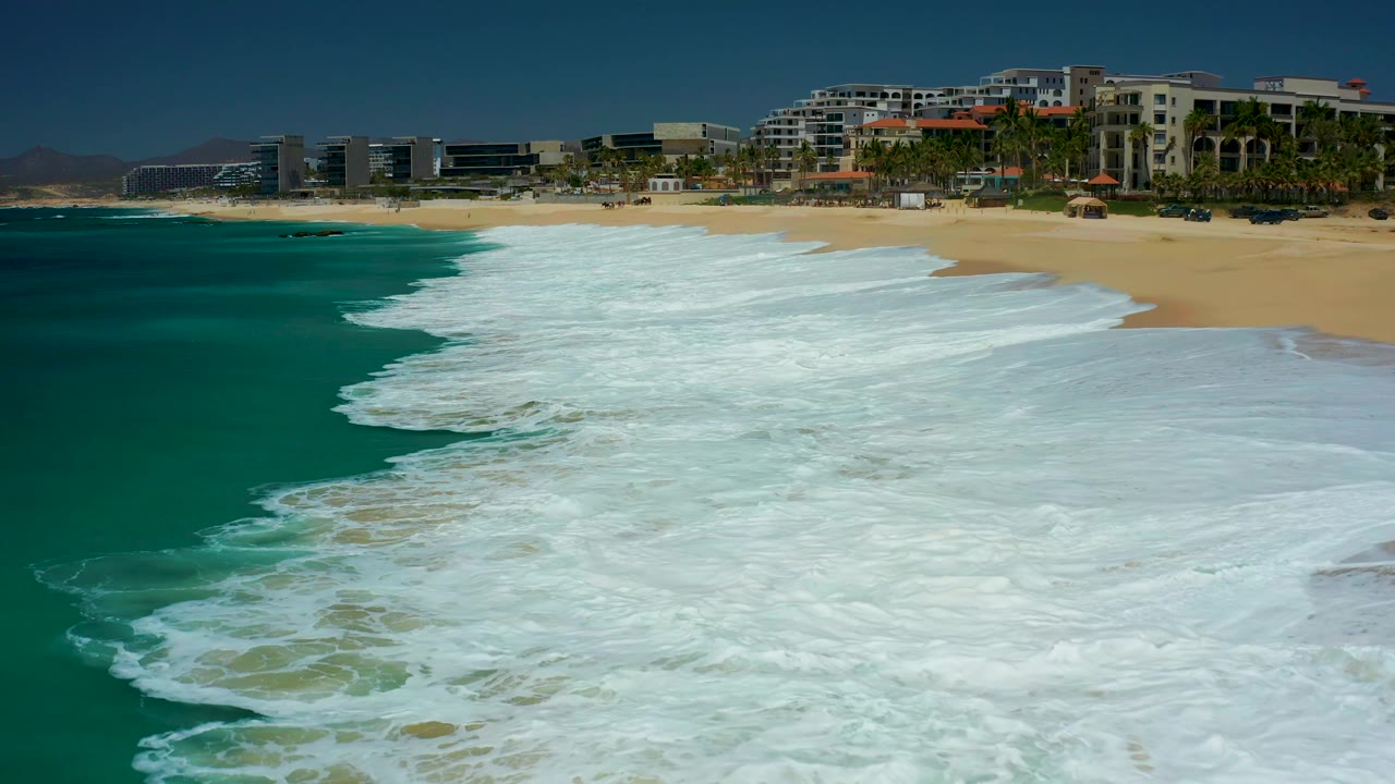 The wave wash covers more of the foreground beach at Dreams Los Cabos, leaving a bright white foam sheet against tan sand, turquoise water, palm trees, and resort buildings, with no visible people.